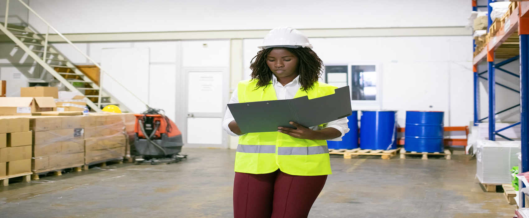 Focused female logistic employee in hardhat and safety vest walking in warehouse, carrying open folder, looking through document. Copy space, front view. Labor and inspection concept